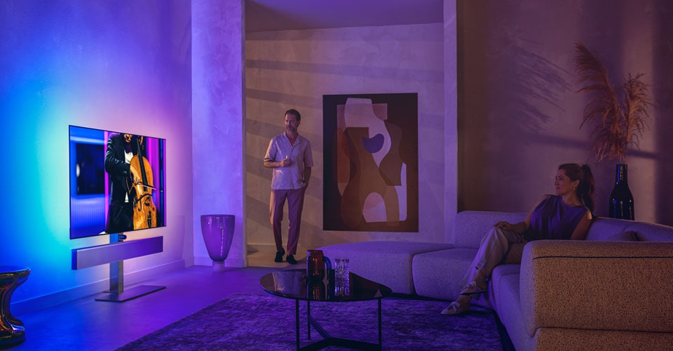 Bărbat și femeie într-un living modern, urmărind un televizor Philips Ambilight care redă un spectacol de violoncel