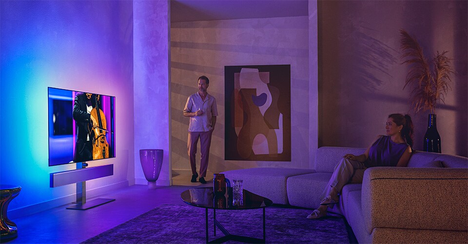 Bărbat și femeie într-un living modern, urmărind un televizor Philips Ambilight care redă un spectacol de violoncel