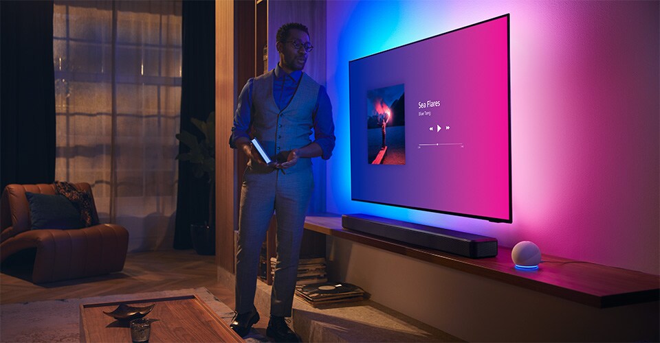 Persoană într-un living modern, lângă un televizor Philips Ambilight cu interfață muzicală și soundbar Fidelio