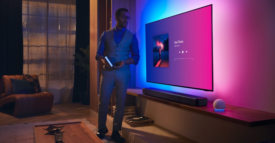 Persoană într-un living modern, lângă un televizor Philips Ambilight cu interfață muzicală și soundbar Fidelio