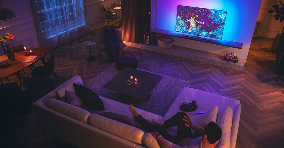 Bărbat relaxându-se pe o canapea într-un living slab luminat, privind un televizor Philips Ambilight