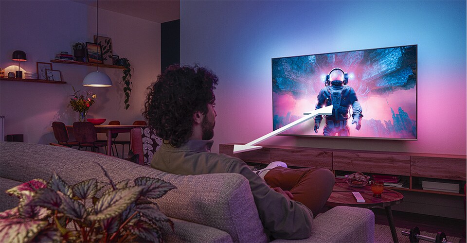 Bărbat așezat pe o canapea, urmărind un televizor Philips Ambilight care afișează o scenă colorată cu un astronaut, cu o săgeată care indică distanța optimă de vizionare