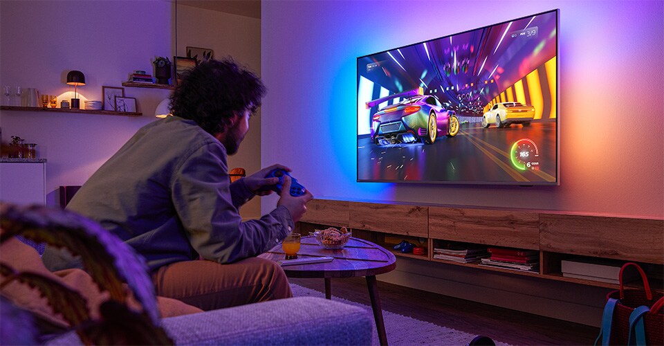 O persoană stă pe canapea și joacă un joc video de curse pe un televizor Philips Ambilight.