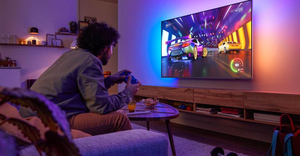 O persoană stă pe canapea și joacă un joc video de curse pe un televizor Philips Ambilight.