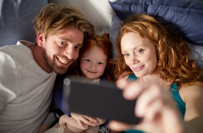 O familie cu un copil mic stau in pat pe niste perne si se pregatesc sa faca o poza in timp ce toata lumea zambeste