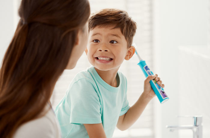 Un băiețel zâmbește larg spre mama lui, în timp ce ține o periuță de dinți Philips Sonicare pentru copii. 